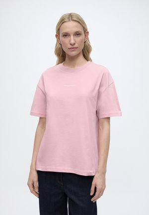 Donna in piedi di fronte, indossa una t-shirt rosa chiaro a maniche corte e jeans blu scuro su uno sfondo bianco uniforme.