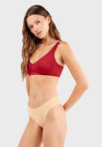 Sostén rojo con tirantes anchos y diseño en forma de V, combinado con unas bragas nude sin costuras que tienen un borde festoneado. Textura suave, estilo sencillo.