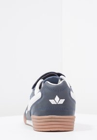 Sneakers navy et blanc avec une semelle en gomme, une sangle Velcro, et un logo géométrique blanc sur le talon, présentés de dos sur une surface blanche.
