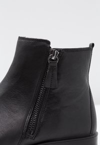 Botín negro de cuero hasta el tobillo con cremallera lateral, textura suave, diseño sin costuras y detalles de costura a lo largo del borde superior.