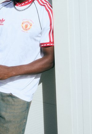 Person trägt weißes Manchester United European Cup Winners 1991 Trikot mit roten Streifen, silberner Kette und hellblauen Jeans, steht an einer Wand.