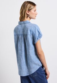 Himmelblau gestreiftes Kurzarmhemd mit abgerundetem Saum und versammeltem Detail auf der Rückseite. Aus einem leichten, strukturierten Stoff gefertigt.