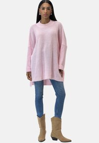 Hellrosa, übergroßer Strickpullover mit einem gerippten Halsausschnitt und seitlichen Schlitzen, kombiniert mit blauen engen Jeans und beigen Cowboy-Stiefeln.