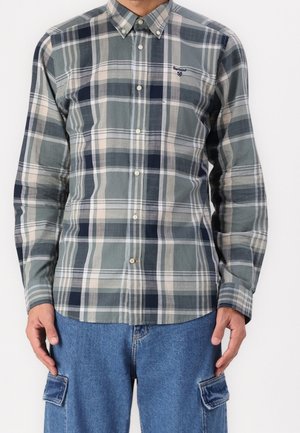 Homme portant une chemise à carreaux verte, beige et bleu marine à manches longues boutonnée et un pantalon cargo en jean bleu.