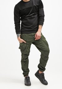 Svart långärmad skjorta med texturerad övre del. Olivgrön cargobyxa med flera fickor. Svarta sneakers med texturerad yta.