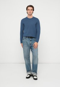 Blå langermet skjorte, lyseblå dongeri jeans, svart belte, svart-hvite sko, stående mot en ensfarget hvit bakgrunn.