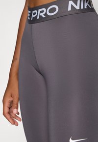 Grijze hoge taille leggings met een gladde textuur, voorzien van een zwarte tailleband met "NIKE PRO" in contrasterend lichtblauw en een wit logo.