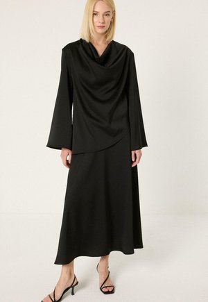 Naine kannab musta pikkade varrukatega kleiti, millel on voltis kaelus ja asümmeetriline hem, mis on kombineeritud mustade rihmikandadega sandaalidega.