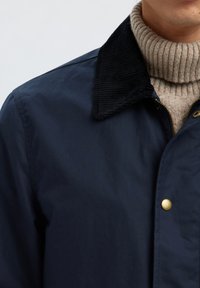 Mies pukeutunut tummansiniseen takkiin, jossa on musta kordurointikaulus ja sen alla oleva beige ribbineuleinen poolopaita.