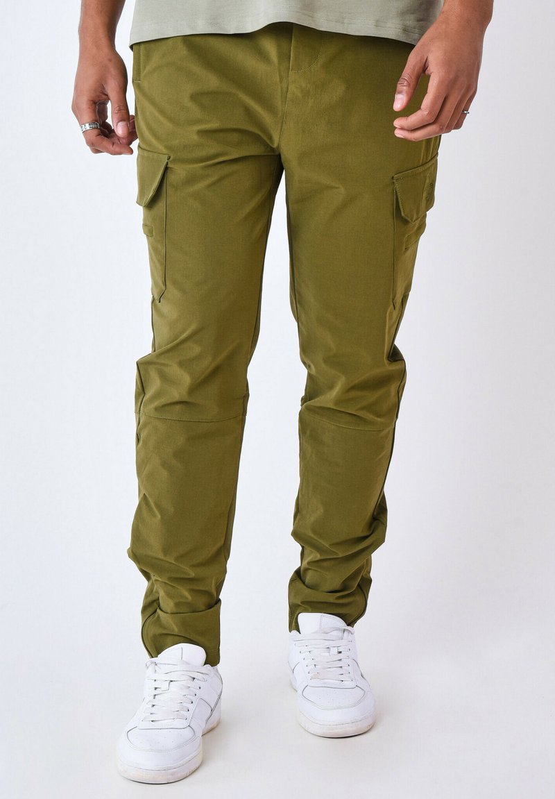 Pantalones cargo verde olivo con bolsillos laterales, piernas ajustadas y una textura suave. Llevados con zapatillas blancas sobre un fondo neutro.
