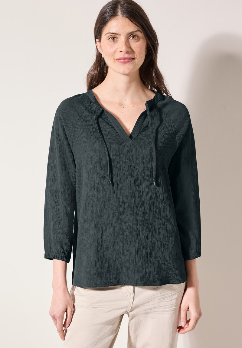 Blusa verde scuro, con texture, a scollo a V e dettaglio con cordino. Presenta maniche a tre quarti e una vestibilità comoda. Perfetta da abbinare a pantaloni di colore chiaro.