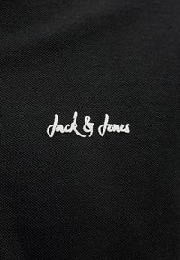 Λευκό λογότυπο "Jack & Jones" με καλλιγραφικά γράμματα σε μαύρο υφασμάτινο φόντο με υφή.