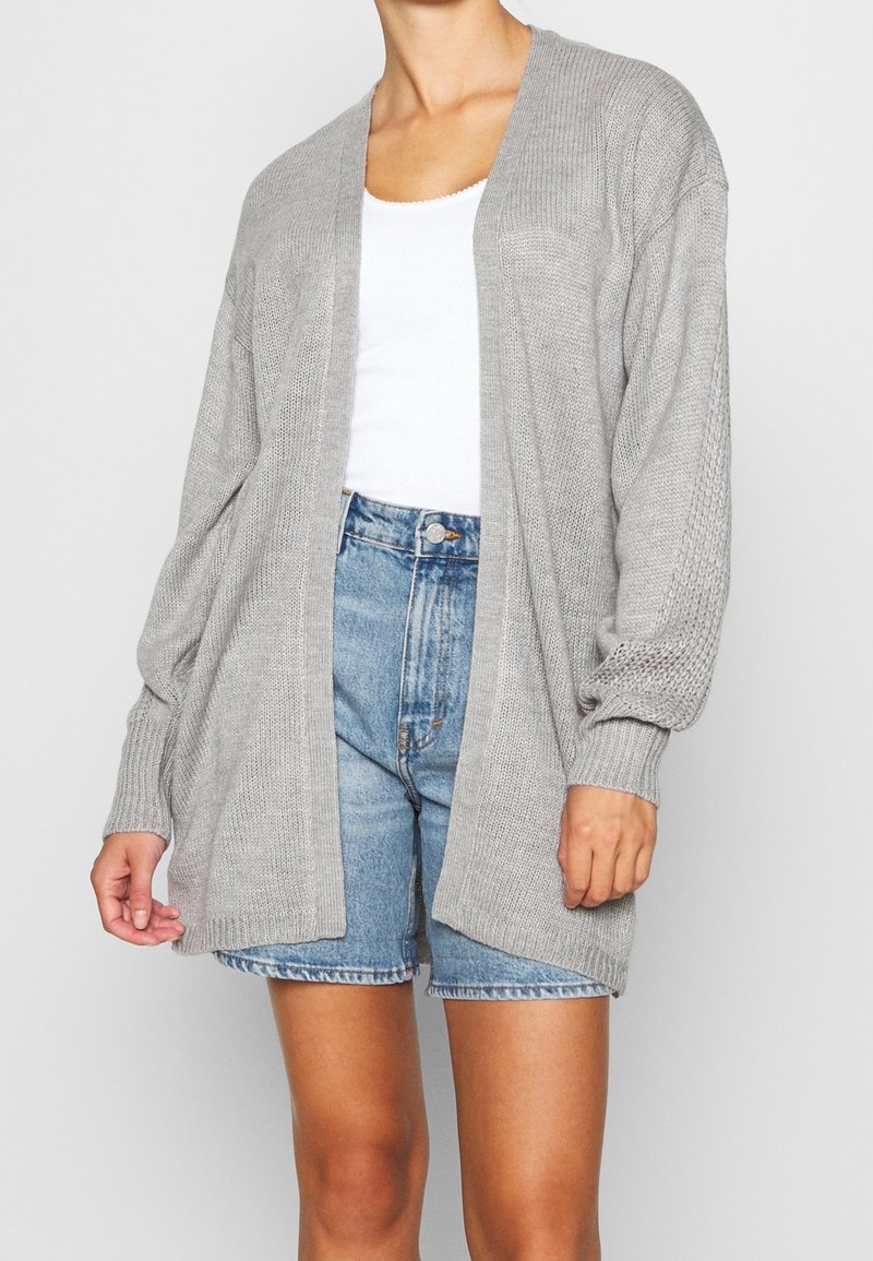 Cardigan aperto grigio chiaro realizzato in morbido tessuto a maglia. Presenta maniche lunghe con polsini a coste e una vestibilità rilassata. Indossato sopra una canotta bianca.
