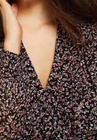 Schwarze Bluse mit floralem Muster und tiefem V-Ausschnitt, verziert mit kleinen lila, rosa und weißen Blumen, Strukturärmel mit elastischen Bündchen und weichem Stoff.