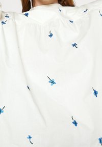 White blouse featuring blue embroidered flowers with green stems, smooth texture, and a gathered design around the neckline.