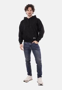 Felpa nera con tasca frontale e coulisse, abbinata a jeans blu medio e sneakers chiare. Il modello è posizionato su uno sfondo bianco.