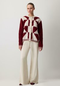 Kardigan z mieszanki wełny z czerwono-kremowym geometrycznym wzorem, z przodem na guziki i prążkowanymi detalami. Połączony z kremowymi spodniami w fasonie dzwonów.