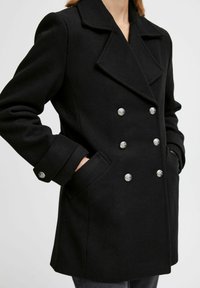 Manteau noir croisé en mélange de laine, avec de grands boutons brillants, des revers larges et des poches latérales, offrant une coupe structurée.