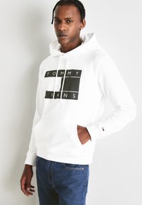 Felpa bianca con cappuccio, caratterizzata da una grande grafica nera con la scritta "TOMMY JEANS." Tasca a marsupio frontale, polsini e orlo a coste, abbinata a jeans blu.