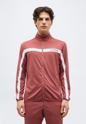 Νεαρός άνδρας με σγουρά σκούρα μαλλιά, φοράει βελούδινο μπορντό αθλητικό μπουφάν με φερμουάρ και λευκές ρίγες, μαζί με παντελόνι που ταιριάζει, μπροστά σε απλό φόντο.