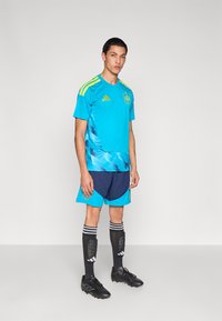 Man wearing blue and navy soccer kit with yellow accents, black shin guards, and black cleats standing against a plain background.