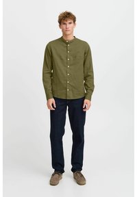Olivgrünes Langarmhemd mit Stehkragen, Knopfverschluss und feiner Textur, kombiniert mit dunkelblauen Jeans und braunen Wildleder-Schuhen.