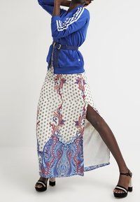 Veste zippée bleue avec des rayures blanches, associée à une jupe maxi blanche ornée d'un motif paisley en rouge et bleu, et d'une fente sur le côté haute jusqu'à la cuisse.