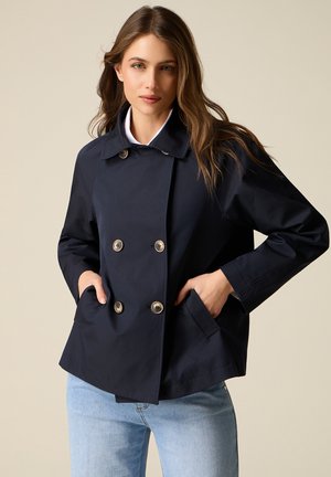 Kvinde iført en mørkeblå dobbeltradet jakke med knappedetaljer og lyseblå jeans, stående mod en ensfarvet beige baggrund.