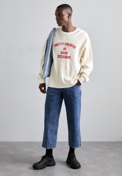 Beige, ribbed sweatshirt with red text; medium-blue denim trousers with a loose fit and white stitching; black shoes; gray jacket draped over shoulder.