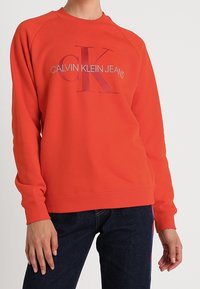 Felpa rossa con collo a giro e maniche raglan, presenta un grande logo "CK" e il testo "CALVIN KLEIN JEANS" in colori a contrasto.