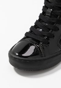 Svart patentlær sneaks med teksturerte svarte lisser og blank tåhette. Har en solid svart gummisåle og sydd detaljer.