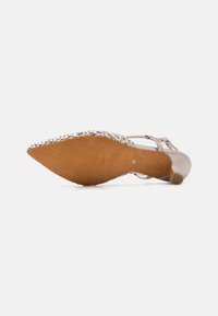 Scarpa con tacco alto con una tomaia sintetica beige, dettagli intrecciati attorno alla punta e una suola marrone testurizzata. Decorazione con fibbia vicino alla caviglia.