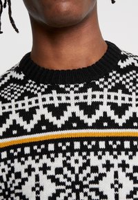 Pull en tricot avec un motif géométrique noir et blanc, col rond et une bande horizontale jaune sur la poitrine.