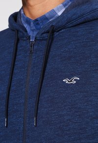 Mörkblå zip-up hoodie i mjukt material, med en känguruficka, snörning i huvan och en vit logotyp på bröstet.