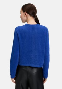 Vrouw met donker, vastgebonden haar die een blauwe, fuzzy trui en zwarte leren broek draagt, met haar rug naar de camera gekeerd tegen een effen lichte achtergrond.