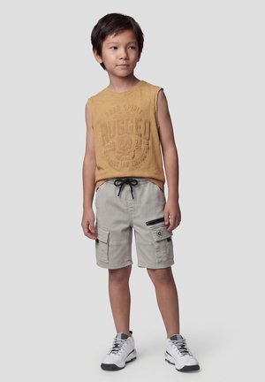 Niño de pie con camiseta sin mangas mostaza con el texto "Rugged", pantalones cortos cargo gris claro con cordón y zapatillas blancas.