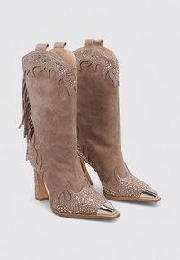 Botas de tobillo de ante marrón con punta metálica en forma puntiaguda, acentos de flecos y patrones decorativos de llamas con tachuelas a lo largo del borde superior.