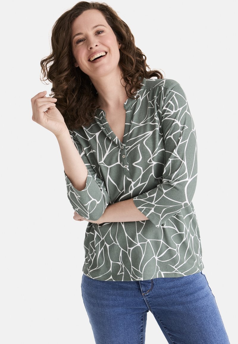 Grün gemusterte Langarmbluse mit einem weißen abstrakten Design und einer knöpfbaren Vorderseite, kombiniert mit blauen Jeans.