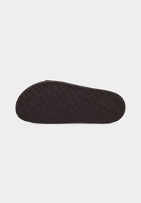Semelle d'une sandale marron foncé avec un motif de bande de roulement géométrique texturé, présentée sur un fond gris clair uni.