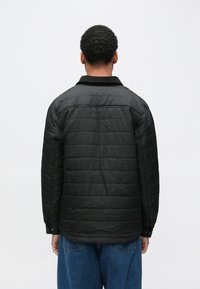 Czarna pikowana kurtka z kołnierzem, widocznymi szwami i guzikami, zestawiona z niebieskimi spodniami dżinsowymi. Widok z tyłu ukazuje fakturę kurtki.