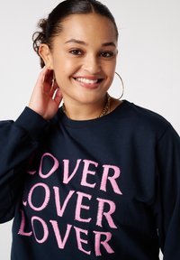 Donkerblauwe sweatshirt met roze geborduurde tekst "LOVER" in een herhaald patroon. Lange mouwen en een relaxed fit.