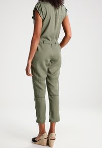 Kobieta ubrana w oliwkowo-zielony kombinezon z paskiem, z podwiniętymi rękawami i beżowymi sandałami na koturnie, stojąca tyłem do widza.