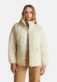 Prošívaná krémová bunda s vysokým límcem, dvěma předními kapsami a elastickými manžetami, se zipovým zapínáním a jemným diamantovým vzorem.