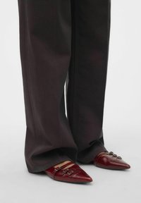 Zapatos rojos de punta afilada con un acabado de cuero brillante y acentos de hebilla, combinados con pantalones negros anchos.