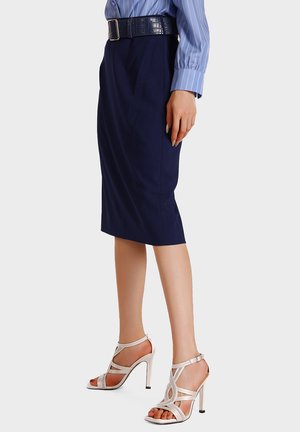 Donna con gonna a matita blu navy, camicia a righe blu, cintura nera e sandali bianchi con tacchi alti, in piedi davanti a uno sfondo semplice.