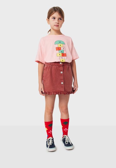 Camiseta de algodón rosa con gráfico de rayuela, falda de pana color óxido con botones en la parte delantera y volantes, calcetines rojos con patrones geométricos y zapatos azul marino.