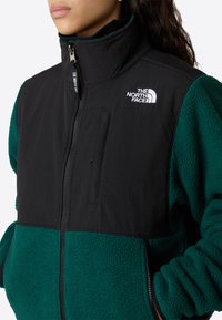 Giacca in pile con parte superiore in nylon nero e parte inferiore verde texturizzata, dotata di zip frontale, colletto alto e logo ricamato sul petto.