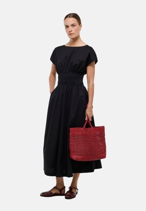 Femme portant une robe midi noire, tenant un grand sac cabas tissé rouge, et portant des sandales fermées marron foncé, debout sur fond blanc.