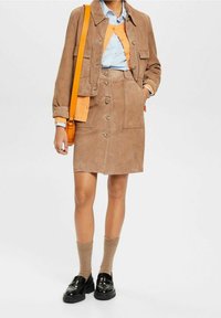 Vrouw in een beige suède kokerrok met knopen en bijpassende jas, lichtblauw shirt, oranje cardigan, beige sokken, zwarte loafers en een oranje schoudertas.