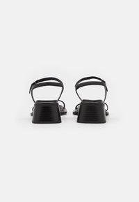 Sandales noires pour femmes avec petits talons carrés et brides chevilles à fermeture par boucle, vues de dos sur fond blanc.